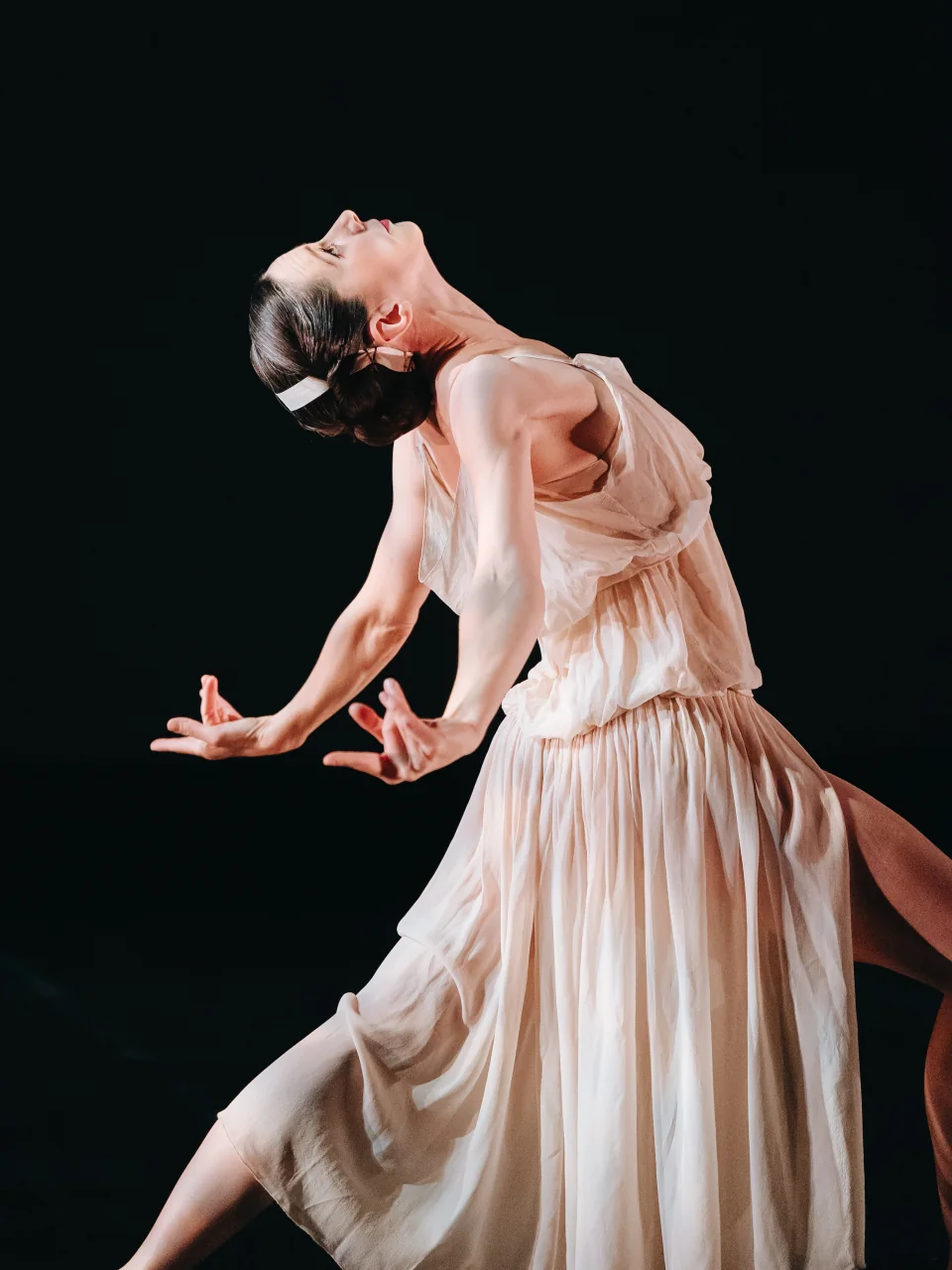 Amy arching backwards with her arms open, wearing a pale dress on a dark stage